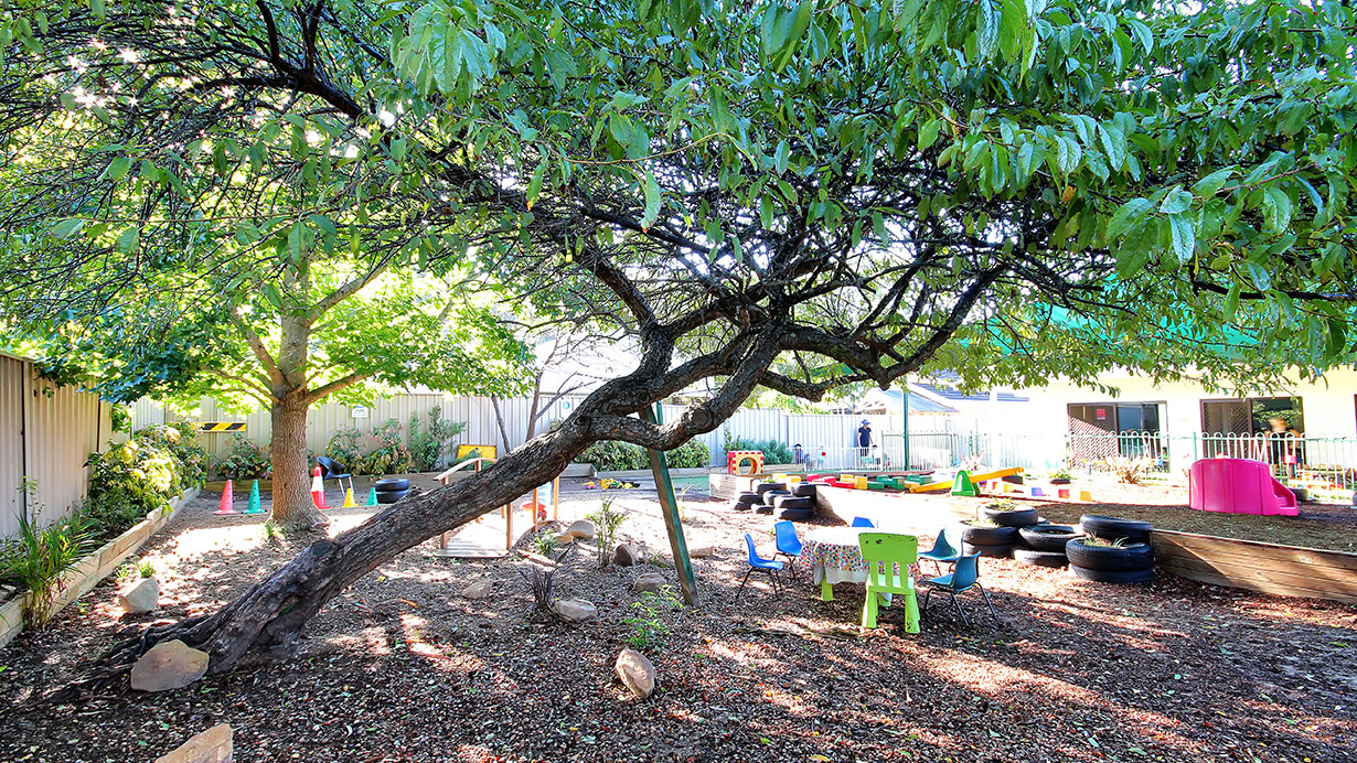 Trees provide shade over childrens bright coloured chairs and sand pit.