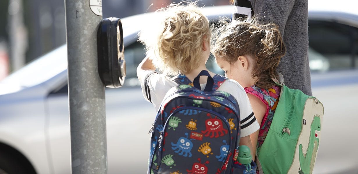 Two children wearing backpacks waiting at traffic light in CBD