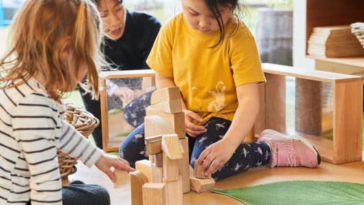 Focused children engage in collaborative block play at Guardian Childcare Springfield, Queensland, building a tower with wooden blocks on a leaf mat, fostering fine motor skills and cooperative learning.