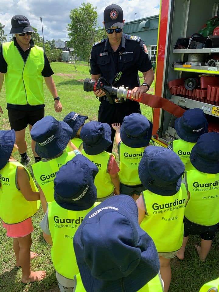 Guardian Brassall children wearing safety vests and hats looking at fire truck hose