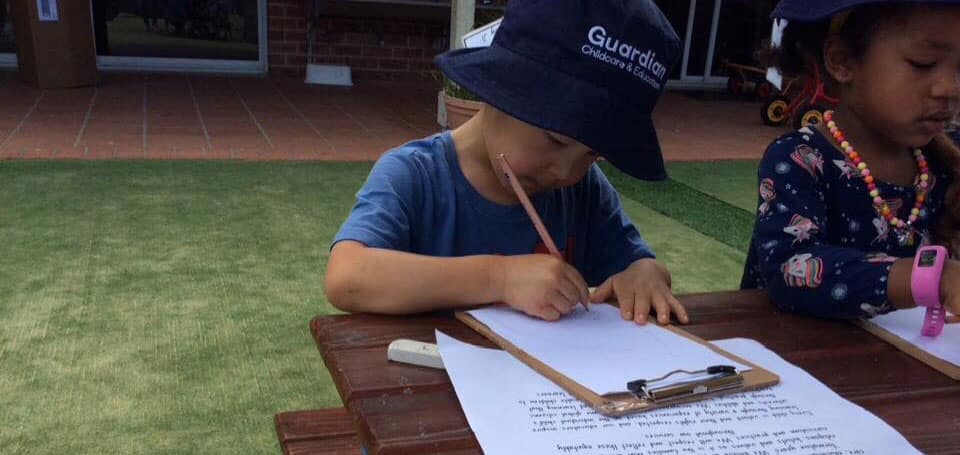 child wearing Guardian hat and writing outside, reviewing centre philosophy