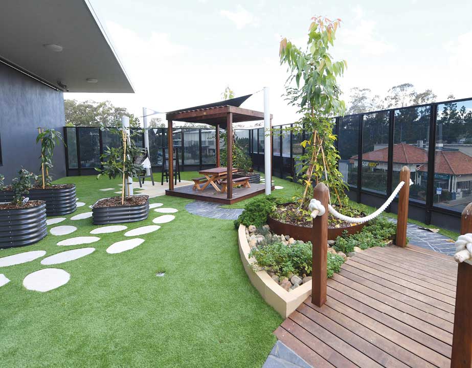 Discover the inviting outdoor play area at Guardian Childcare Springfield Lakes, featuring a shaded pergola, nature play garden beds, and safe artificial turf.