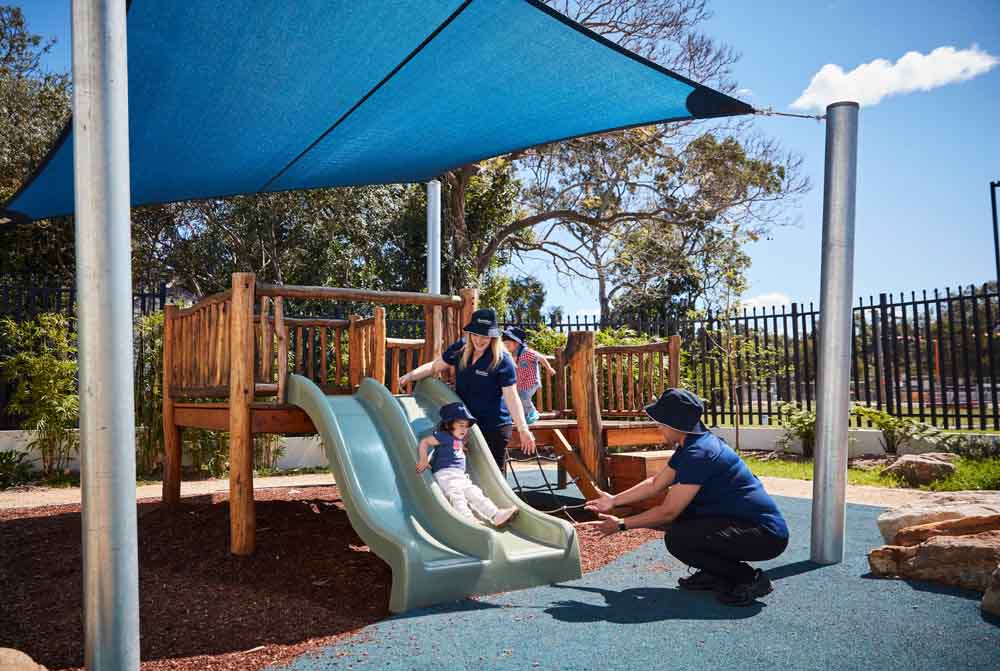 Children enjoying outdoor play on a wooden slide and play structure at Guardian Childcare Parramatta, under a bright blue shade sail.