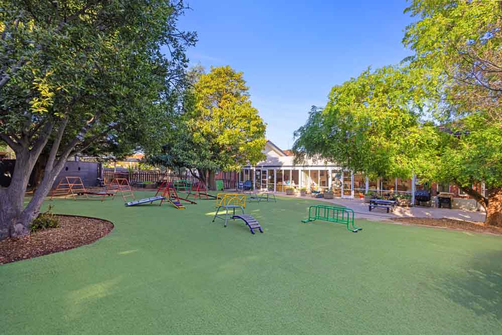 Vibrant outdoor playground at Guardian Childcare Camberwell VIC, featuring artificial grass, diverse play equipment including slides, climbing frames, and balance beams, under a sunny sky.