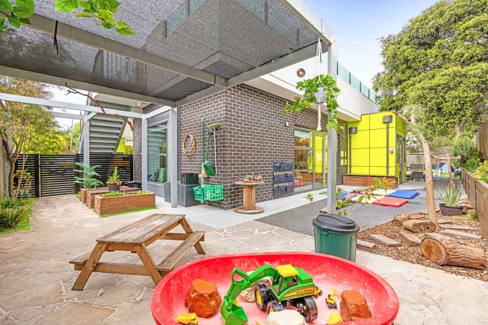 Outdoor play area at Guardian Childcare Blackburn South with a sandpit filled with toy trucks and a picnic table under shade.