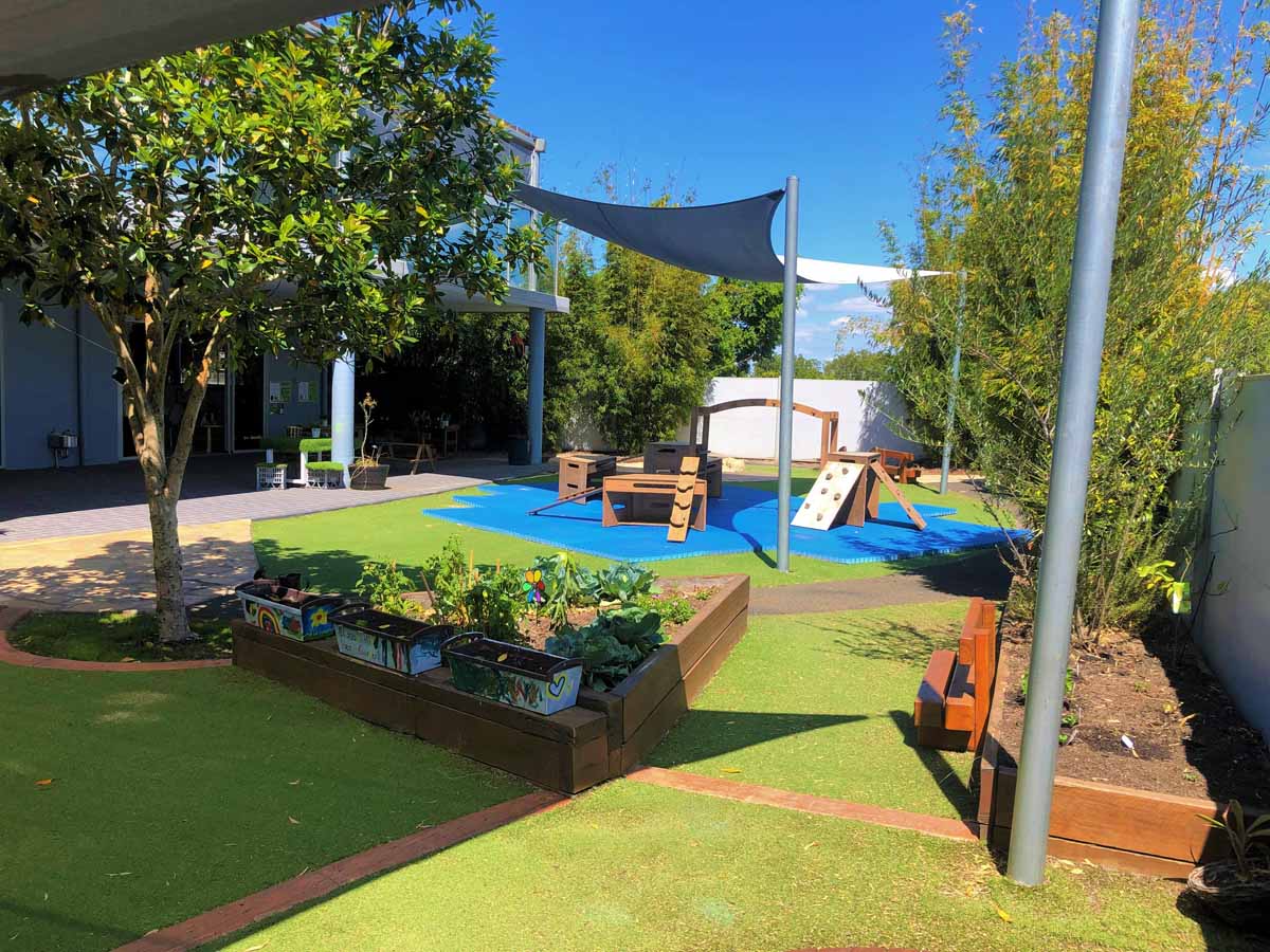 Extensive shaded outdoor play space at Guardian Childcare Hamilton QLD, featuring natural wooden climbing frames, garden beds for nature play, and safe artificial turf.