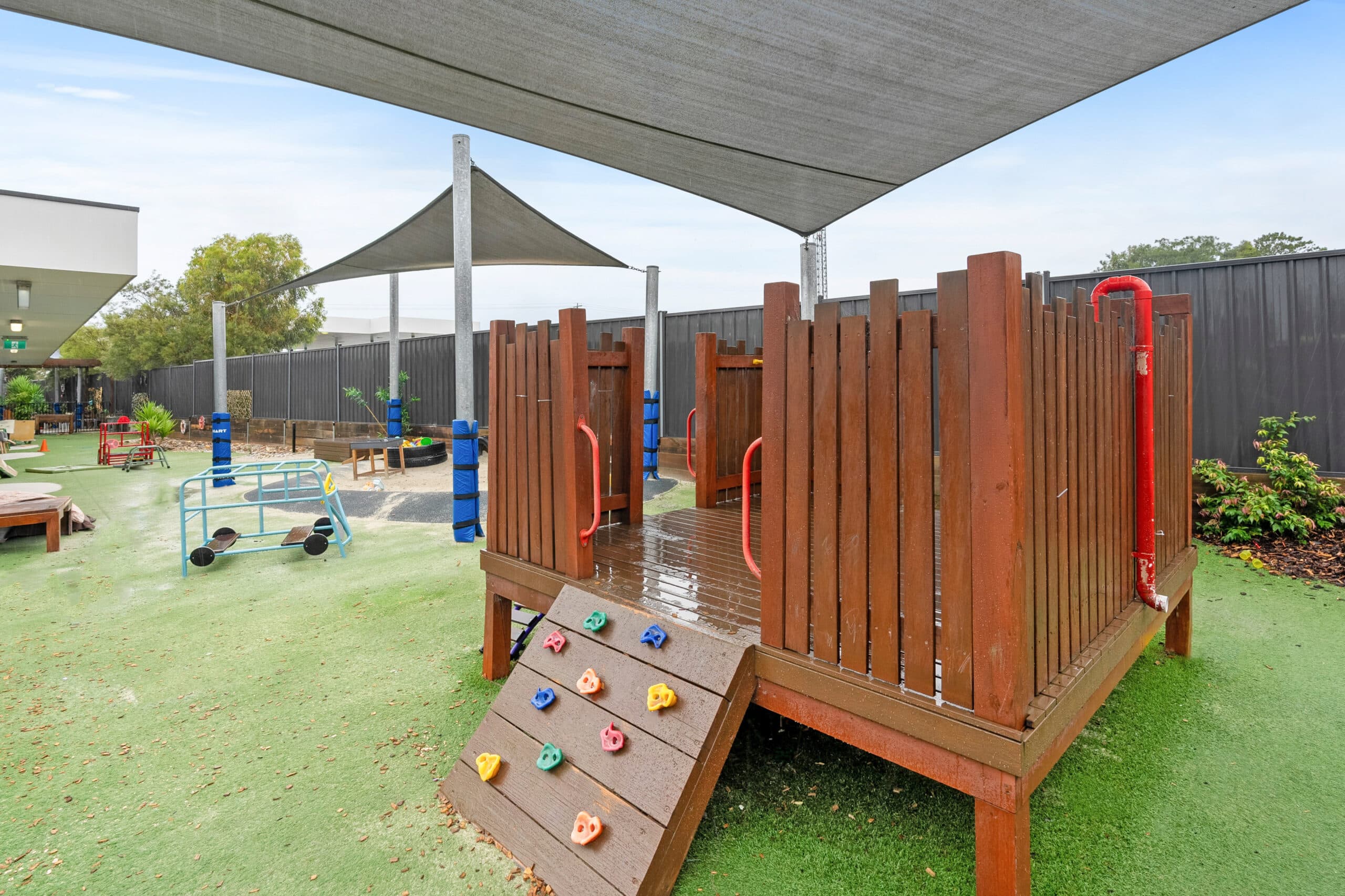 Covered wooden play structure with climbing wall at Guardian Childcare Leopold, Victoria. Features colourful handholds, ramp access, artificial grass safety surface, and shade protection.